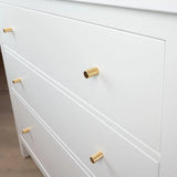 Een close-up van een witte commode met drie lades, elk voorzien van een Remo Gold Deluxe knop van yessential.nl. De cilindervormige gouden handgrepen brengen een vleugje elegantie als het dressoir op een lichte houten vloer staat.
