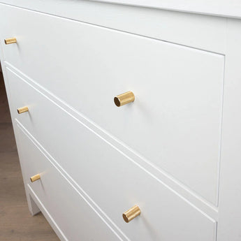 Een close-up van een witte commode met drie lades, elk voorzien van een Remo Gold Deluxe knop van yessential.nl. De cilindervormige gouden handgrepen brengen een vleugje elegantie als het dressoir op een lichte houten vloer staat.