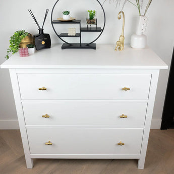 Een moderne witte ladekast met drie lades en de Arturo Gold knoppen van yessential.nl is versierd met planten, decoratieve accenten, een zwarte plank en een witte vaas, die landelijke interieur charme brengt aan de lichte muur en vloer.