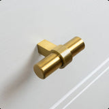 Een close-up van de Arturo Gold Knop van yessential.nl, met een mat gouden T-vormig ontwerp gemonteerd op een lade met witte panelen - een ideale keuze voor een landelijk interieur.