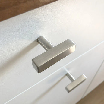 Close-up van een moderne geborsteld metalen Vasco RVS knop van yessential.nl, gemonteerd op een wit ladefront met een houten aanrecht zichtbaar aan de bovenrand.