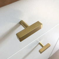 Een close-up van twee moderne Vasco Gold Knobs van yessential.nl, met een luxe gouden afwerking en geborstelde afwerking, verticaal gemonteerd op een wit ladefront.