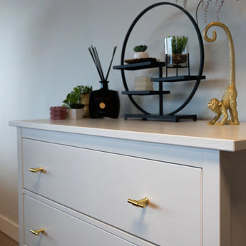 Het Cordero Gold dressoir van yessential.nl heeft gouden handgrepen en een deurknop goud, gestyled met vetplanten in potten, een rietverdamper, een ronde zwarte plank, kaarsen en een gouden apenornament met een krulstaart.