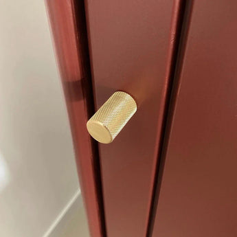 Een close-up van de Remo Gold Deluxe knop van yessential.nl, een luxe meubelknop goud met een gestructureerd cilindrisch ontwerp, getoond op een mat donkerrode kast of lade.