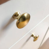 Close-up van twee 10x Antoni Matte Gold kastknoppen van yessential.nl op een witte kast, gefotografeerd onder een hoek om de gladde, glanzende afwerking te laten zien.