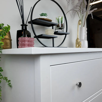 Een witte commode met yessential.nl Marizo RVS meubelknoppen en een ronde zwarte plank met kleine plantjes en decor. Op het dressoir staan een roze noppenkaars, een vaas, groen en een gouden apenbeeldje. Op de achtergrond staan nog meer planten en een spiegel.