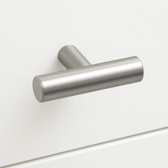 Een Remo RVS knop van yessential.nl, met een geborsteld metalen T-vorm en mat cilindrisch ontwerp, is gemonteerd op een witte kastlade voor een eenvoudige, moderne look.