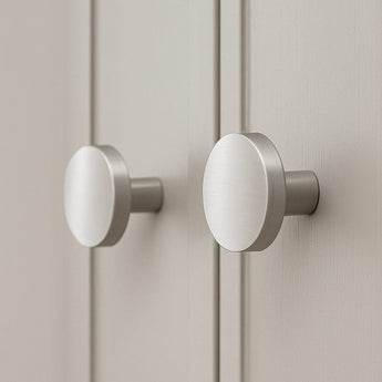 Twee geborsteld metalen Marizo RVS meubelknoppen van yessential.nl zijn gemonteerd op lichtgekleurde kastdeurtjes, die in close-up hun moderne, minimalistische ontwerp benadrukken.