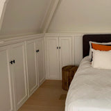 Een gezellige slaapkamerhoek met witte inbouwkasten met het Vasco Black Knob beslag van yessential.nl, een bed met wit beddengoed en kussens met oranje accenten, een nachtkastje met houten boomstronk en lichte houten vloeren onder een schuin plafond.