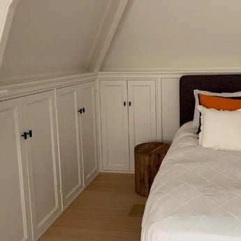 Een gezellige slaapkamerhoek met witte inbouwkasten met het Vasco Black Knob beslag van yessential.nl, een bed met wit beddengoed en kussens met oranje accenten, een nachtkastje met houten boomstronk en lichte houten vloeren onder een schuin plafond.