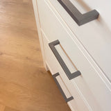 Close-up van een witte kast met drie lades, elk voorzien van Vasco Gunmetal handgrepen van yessential.nl. Geplaatst op een lichte houten vloer combineert dit meubel moderne stijl en functionaliteit voor elke kamer.