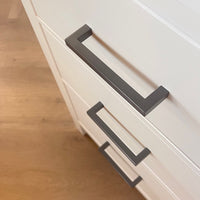 Close-up van drie witte lades met moderne Vasco Gunmetal handgrepen van yessential.nl, tegen een lichte houten vloer. De lades hebben een lichte hoek, waardoor de strakke lijnen en het minimalistische ontwerp goed tot hun recht komen.
