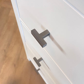 Close-up van twee Vigio Gunmetal knoppen van yessential.nl op witte kastladen met een houten vloer op de achtergrond, voor een vleugje moderne meubelknop stijl.