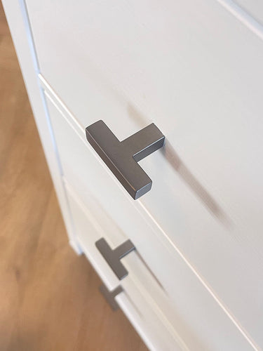 Close-up van twee Vigio Gunmetal knoppen van yessential.nl op witte kastladen met een houten vloer op de achtergrond, voor een vleugje moderne meubelknop stijl.