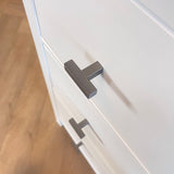Close-up van twee Vigio Gunmetal knoppen van yessential.nl op witte kastladen met een houten vloer op de achtergrond, voor een vleugje moderne meubelknop stijl.
