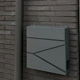 De Sergio Anthracite brievenbus van yessential.nl, met strakke lijnen, is bevestigd op een donkere bakstenen muur naast een bijpassende grijze regenpijp.