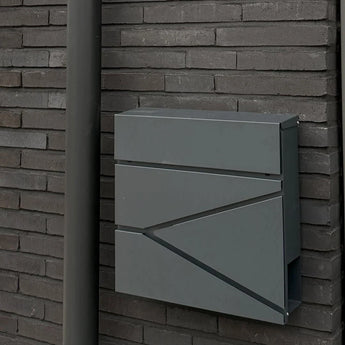 De Sergio Anthracite brievenbus van yessential.nl, met strakke lijnen, is bevestigd op een donkere bakstenen muur naast een bijpassende grijze regenpijp.