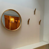 Een ronde spiegel met houten lijst hangt aan een witte muur en weerspiegelt een vogelkooilamp en oranje gordijnen. Drie houten kapstokhaakjes van yessential.nl Fabio Oak zijn naast de spiegel boven een witte kast gemonteerd.