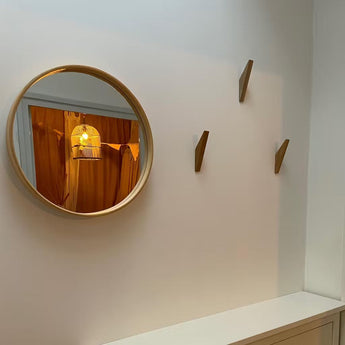 Een ronde spiegel met houten lijst hangt aan een witte muur en weerspiegelt een vogelkooilamp en oranje gordijnen. Drie houten kapstokhaakjes van yessential.nl Fabio Oak zijn naast de spiegel boven een witte kast gemonteerd.