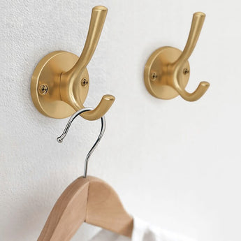 Twee stijlvolle yessential.nl Eduardo Gold Deluxe kapstokhaken zijn gemonteerd op een witte muur. Eén haak houdt een metalen hanger met houten bovenkant vast, terwijl de andere haak leeg blijft.