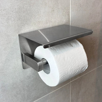 Een rol toiletpapier op een yessential.nl Tazio RVS toiletrolhouder met planchet, gemonteerd op een lichtgrijs betegelde badkamermuur.