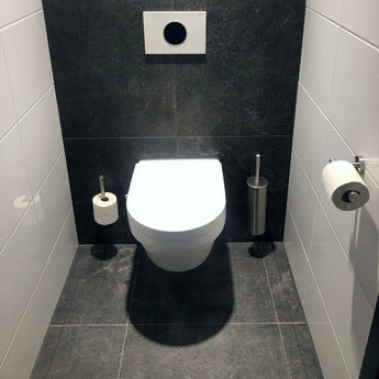 Een moderne badkamer met links een wandcloset, de Tino RVS toiletrolhouder van yessential.nl, rechts toiletaccessoires zoals een borstel, daarboven een doorspoelknop en grijs-witte tegels op zowel de vloer als de muren tegen donkere accenten.