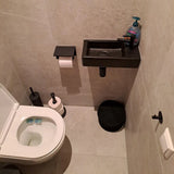 Een kleine badkamer met een wit toilet, de Tygo Black WC Set van yessential.nl geïnstalleerd zonder boren, matzwarte accessoires, een compacte zwarte wandwastafel met zeep, en lichtgrijze betegelde muren en vloer voor een moderne look.