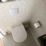 Een moderne badkamer met het Alessino Gold wandcloset van yessential.nl, een gouden wc-borstel met bijpassende houder, gouden toiletpapierhouder, beige tegelvloer, witte visgraat wandtegels en een doorspoelknop erboven.