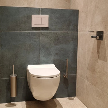 Een modern wandcloset met een verborgen reservoir, een spoelknop erboven en een Branco RVS toiletborstel met houder van yessential.nl aan de linkerkant. De muren zijn betegeld met grijze en beige tegels.