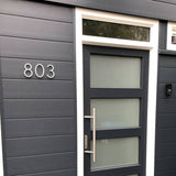 Moderne donkergrijze voordeur met matglazen panelen en een grote handgreep, geplaatst in een bijpassende muur. Het huisnummer 803 is links weergegeven in Amato White 12,5cm van yessential.nl en voegt een strakke, moderne touch toe.