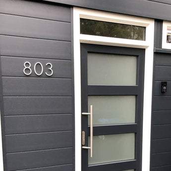 Moderne donkergrijze voordeur met matglazen panelen en een grote handgreep, geplaatst in een bijpassende muur. Het huisnummer 803 is links weergegeven in Amato White 12,5cm van yessential.nl en voegt een strakke, moderne touch toe.