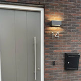 Een moderne grijze deur met een lange verticale handgreep staat bij een bakstenen muur met het Amato White 12,5cm huisnummer van yessential.nl, een zwarte brievenbus, deurbelknop en een verlichte wandlamp boven het nummer.