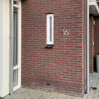 Een rood bakstenen huis met een smal verticaal raam en moderne huisnummers. Het nummer 16, Amato White 15cm  is op de muur bevestigd naast een grijze regenpijp, met geplaveide stenen op de grond.