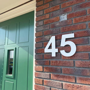 Een close-up van een bakstenen muur toont het yessential.nl Adamo White 15cm huisnummer "45" naast een groene voordeur met een rechthoekig raam, zonlicht werpt schaduwen over beide.