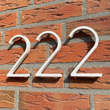 Amato White 12,5cm huisnummers van yessential.nl in "wit 222" zijn gemonteerd op een rode bakstenen muur met grijze mortel, die opvallende schaduwen werpt in het zonlicht voor een moderne look.