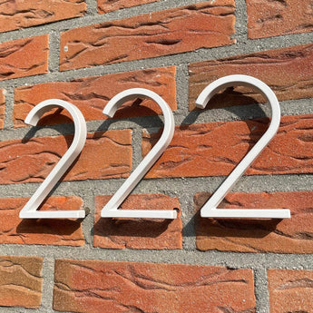 Amato White 12,5cm huisnummers van yessential.nl in "wit 222" zijn gemonteerd op een rode bakstenen muur met grijze mortel, die opvallende schaduwen werpt in het zonlicht voor een moderne look.