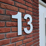 Het Adamo White 30cm huisnummer van yessential.nl, in maat 13, wordt gemonteerd op een rode bakstenen muur naast een wit omlijst raam of deur.