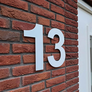 Het Adamo White 30cm huisnummer van yessential.nl, in maat 13, wordt gemonteerd op een rode bakstenen muur naast een wit omlijst raam of deur.