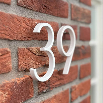 De Amato White 12,5cm huisnummers van yessential.nl zijn gemonteerd op een rode bakstenen muur, gefotografeerd onder een hoek. De moderne, verhoogde cijfers werpen een schaduw, met rechts een deel van een raamkozijn zichtbaar.