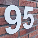 De Adamo White 20cm huisnummer 95 van yessential.nl steekt mooi af tegen een rode bakstenen muur, waardoor gebouw- of huisnummers goed zichtbaar zijn.