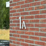 Amato Witte 20cm huisnummers van yessential.nl zijn gemonteerd op een rode bakstenen muur, in de stijl van moderne huisnummers. Wat groen en een deel van een zwarte auto verschijnen vaag op de achtergrond.