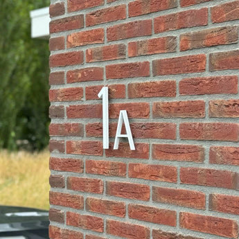 Amato Witte 20cm huisnummers van yessential.nl zijn gemonteerd op een rode bakstenen muur, in de stijl van moderne huisnummers. Wat groen en een deel van een zwarte auto verschijnen vaag op de achtergrond.