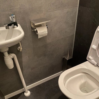 Een kleine badkamer met een witte wastafel, een open wit toilet en donkergrijze betegelde muren en vloer, met de yessential.nl Tazio Chrome (No Drill) toiletrolhouder voor gemakkelijke toegang tot toiletpapier.