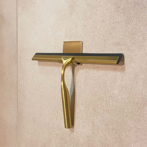 De Zamoro Gold van yessential.nl, met een goudkleurige douchetrekker met een zwart rubberen blad, is gemonteerd op een beige betegelde muur in een bijpassende gouden houder.