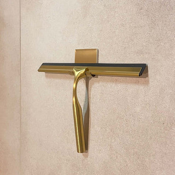 De Zamoro Gold van yessential.nl, met een goudkleurige douchetrekker met een zwart rubberen blad, is gemonteerd op een beige betegelde muur in een bijpassende gouden houder.