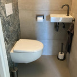 Een kleine, moderne badkamer heeft een toilet aan de muur, een compacte witte wastafel met een handdoek eronder en de Tygo Gunmetal WC Set (no-drill) van yessential.nl, inclusief een toiletpapierhouder en reserverol aan de muur naast de wastafel.